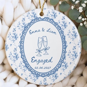 May include: A round white ceramic ornament featuring a blue floral pattern and a central oval lace border. The text reads Emma & Liam, Engaged, and 02.06.2027, with a graphic of two champagne flutes. A gold string is attached to the top for hanging.