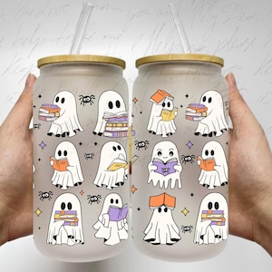 May include: Two clear glass tumblers with a frosted finish and a straw. The tumblers are decorated with a repeating pattern of ghosts reading books. The ghosts are white with purple accents and are surrounded by stars and spiders.