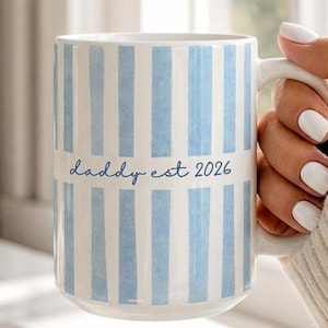 May include: A white ceramic mug with vertical light blue stripes and the text "daddy est 2026". The mug is held by a person with white nail polish. The mug is near a vase of white flowers.