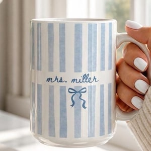 May include: A white ceramic mug with vertical light blue stripes. The mug features the text "mrs. miller" in a script font, along with a blue bow illustration. The mug is held by a person.