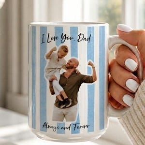 May include: White ceramic mug with blue vertical stripes and a photo of a father and child. The mug has the text "I Love You, Dad" and "Always and Forever". The mug is held by a person.