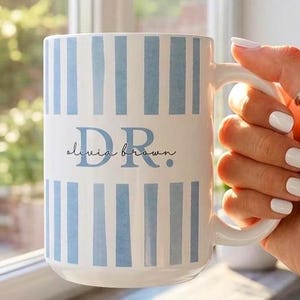 May include: White ceramic mug with blue vertical stripes and the text "DR. olivia brown" in a script font. The mug is held by a hand with white nail polish, a silver ring, and is set against a window.