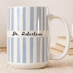 May include: A white ceramic mug with vertical pale blue stripes. The mug has a large handle and the text "Dr. Robertson" in black script. The mug is on a beige woven placemat.