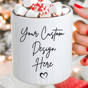 May include: A white ceramic mug filled with hot chocolate, topped with marshmallows and red and white sprinkles. The mug has the text "Your Custom Design Here" in black script with a heart symbol. The mug is held by a person with red nail polish.