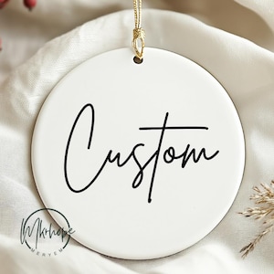May include: A white, circular ceramic Christmas ornament with the word "Custom" in black script. It hangs from a gold string. A small logo is at the bottom left. The background is a soft, white fabric.