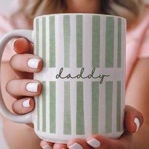 May include: White ceramic mug with a green striped design and the word "daddy" in a handwritten font. The mug has a large handle. The mug is held up in front of a person.