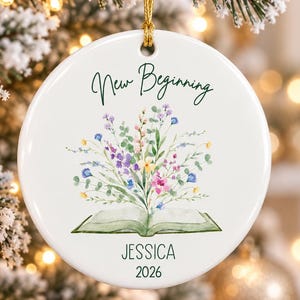 May include: A round white ceramic ornament featuring a watercolour illustration of an open book with a bouquet of colourful wildflowers emerging from the pages. The text New Beginning is printed above, with the name JESSICA and the year 2026 below.