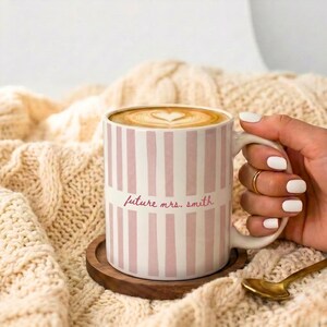May include: A white ceramic mug with pink vertical stripes and the text "future mrs. Smith". The mug is filled with coffee, topped with a heart-shaped design, and rests on a wooden coaster.