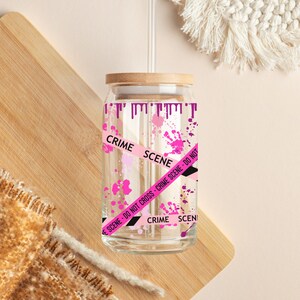 Puede incluir: Un vaso de vidrio transparente con tapa de bamb&uacute; y un dise&ntilde;o rosa y negro de "Escena del crimen - No cruzar". El vaso tiene salpicaduras y gotas de pintura rosa.