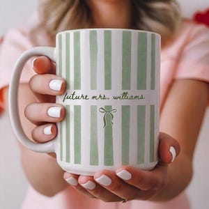 May include: White ceramic mug with vertical green stripes and the text "future mrs. williams" in a script font. A green bow graphic is below the text. The mug has a large handle.