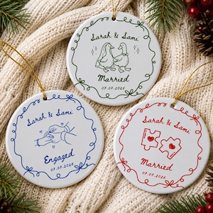 May include: Three round white ceramic ornaments with gold hanging strings. One features green line art of two ducks, the text Sarah & Sami, Married, and 07.07.2026. Another shows blue line art of hands exchanging rings, Sarah & Sami, Engaged, and 07.07.2026. The third has red line art of two puzzle pieces with hearts, Sarah & Sami, Married, and 07.07.2026.