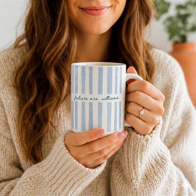 May include: A white ceramic mug with vertical light blue stripes. The mug has the text "future mrs. williams" in a cursive font. The mug is held by a person wearing a beige sweater.