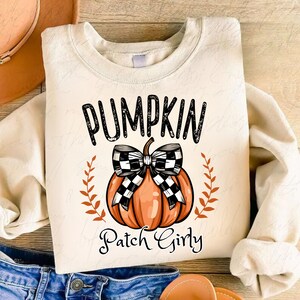 K&ouml;nnte beinhalten: Ein cremefarbener Pullover mit dem Text "Pumpkin Patch Girly" in schwarzen Buchstaben. Eine schwarz-wei&szlig; karierte Schleife befindet sich auf einem orangefarbenen K&uuml;rbis.