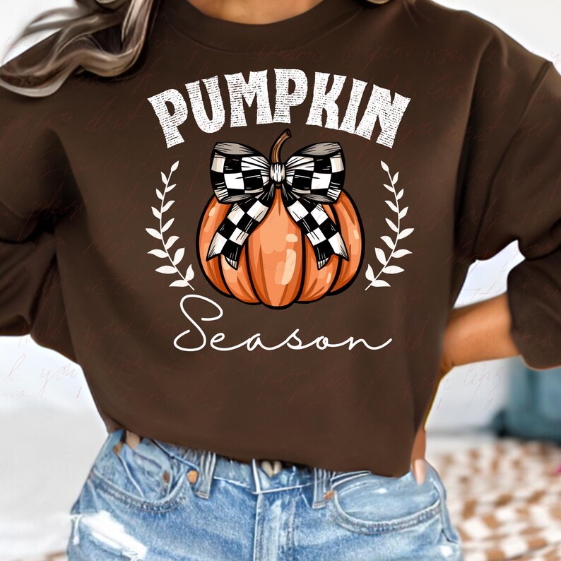 May include: A brown sweatshirt with the text "Pumpkin Season" in white lettering. A cartoon pumpkin with a black and white checkered bow is centered on the sweatshirt.