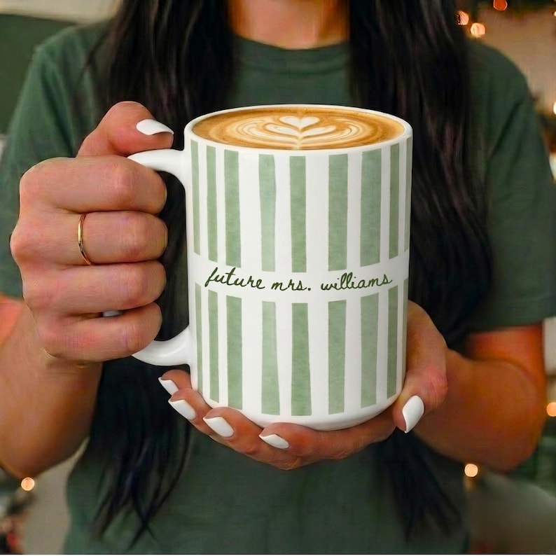 May include: A white ceramic mug with vertical green stripes, filled with coffee and latte art. The mug has the text "future mrs. williams" in black script. The mug is held by a person.