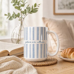 May include: A white ceramic mug with vertical blue stripes and the text "future mrs. williams" in a script font. The mug sits on a round, woven coaster. A croissant rests on a small white plate.