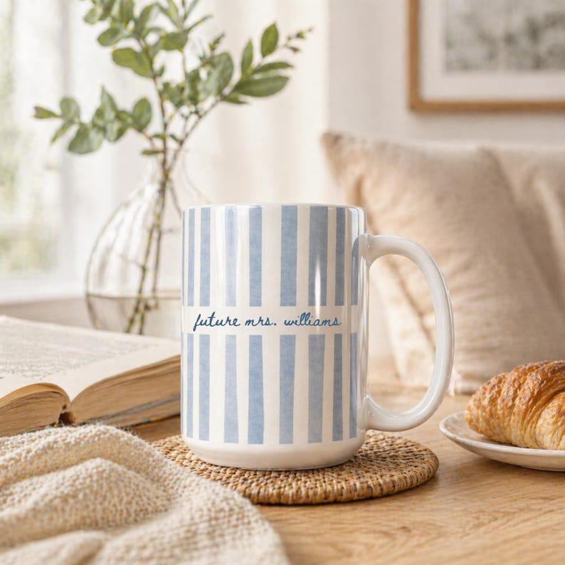 May include: A white ceramic mug with vertical blue stripes and the text "future mrs. williams" in a script font. The mug sits on a round, woven coaster. A croissant rests on a small white plate.