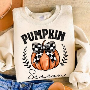 May include: A cream-colored sweatshirt with the text "Pumpkin Season" in black lettering. A black and white checkered bow is tied around an orange pumpkin.