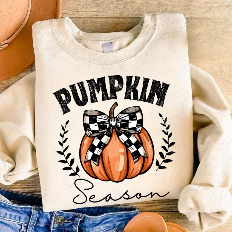 May include: A cream-colored sweatshirt with the text "Pumpkin Season" in black lettering. A black and white checkered bow is tied around an orange pumpkin.
