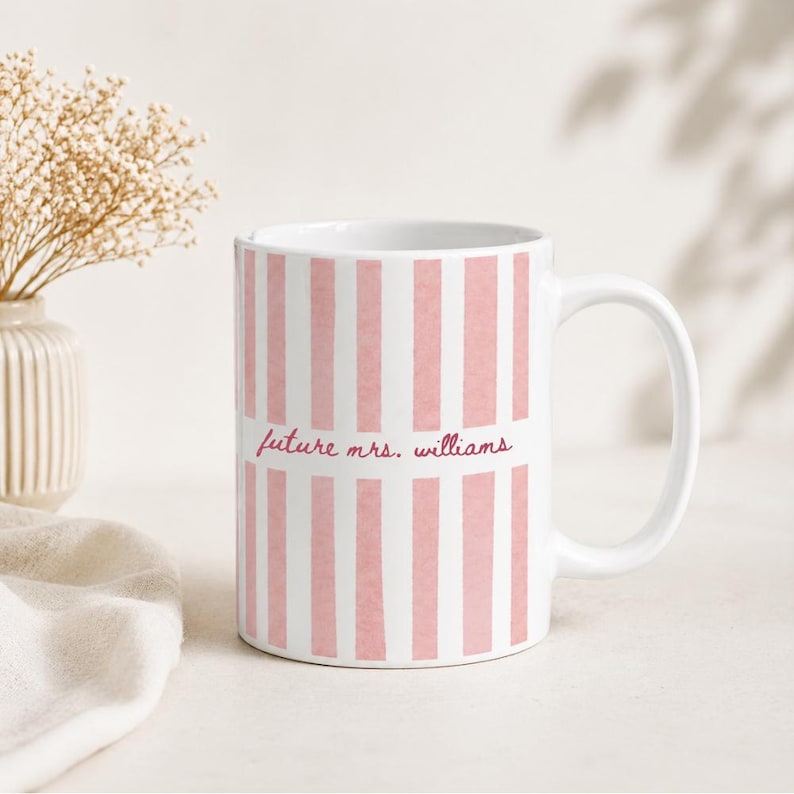 May include: White ceramic mug with vertical pink stripes. The mug features the text "future mrs. williams" in a cursive font. The mug has a curved handle and is likely used for beverages.