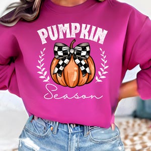 May include: A pink sweatshirt with a white graphic of a pumpkin with a black and white checkered bow. The text "Pumpkin Season" is written below the pumpkin.