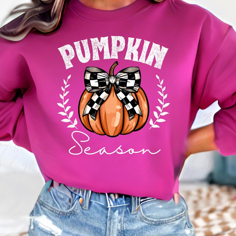 May include: A pink sweatshirt with a white graphic of a pumpkin with a black and white checkered bow. The text "Pumpkin Season" is written below the pumpkin.