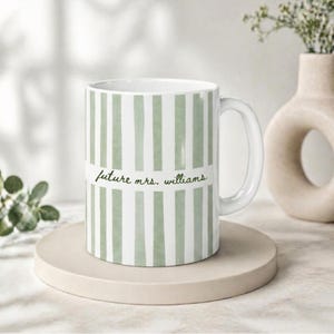 May include: White ceramic mug with vertical green stripes. The mug features the text "future mrs. williams" in a handwritten style. The mug is on a beige circular coaster.