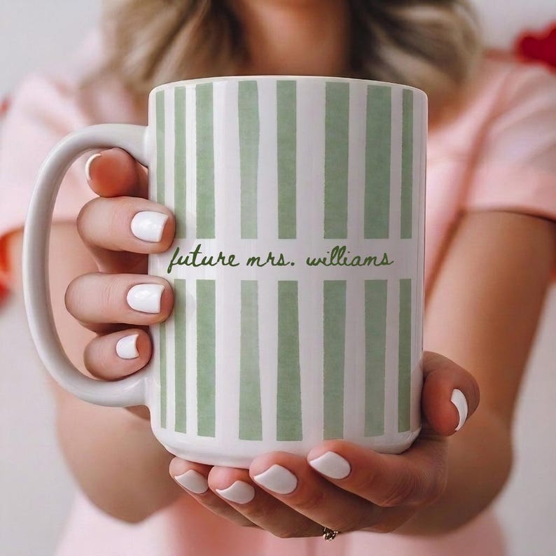 May include: White ceramic mug with a green striped pattern and the text "future mrs. williams" in a script font. The mug has a large handle and is held by a person with white nail polish.