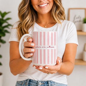 May include: A large white mug with vertical pink stripes. The mug has a large handle and the words "future mrs. williams" in a cursive font. The mug is held by a person wearing a white t-shirt.