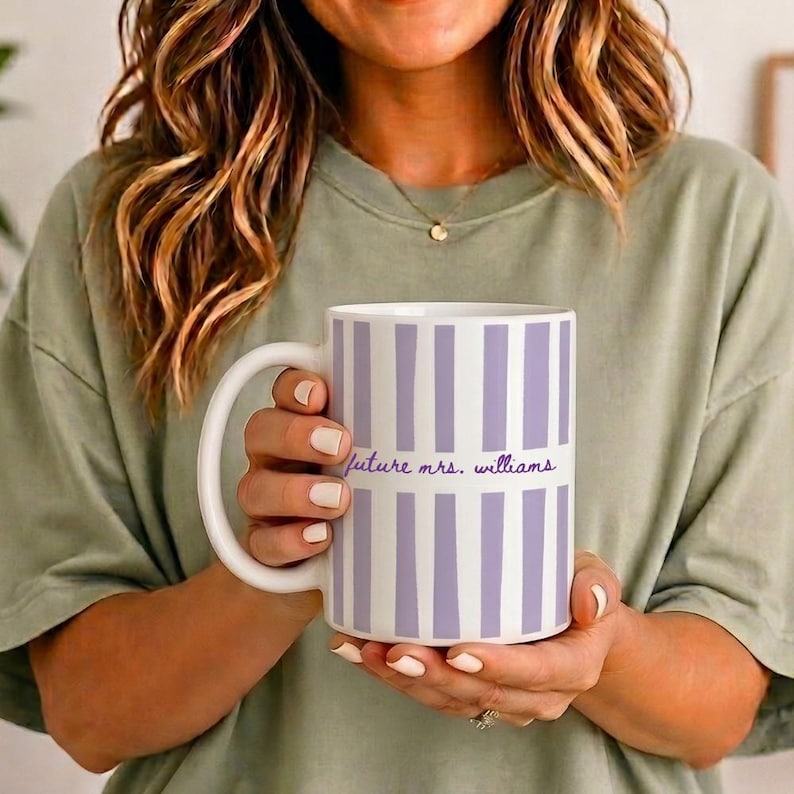 May include: A white ceramic mug with vertical lavender stripes and the text "future mrs. williams" in a script font. The mug has a large, curved handle and is held by a person.