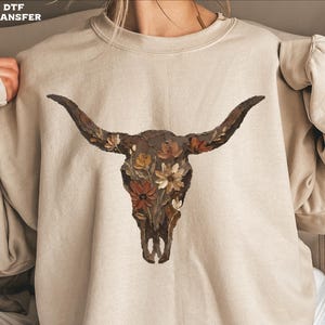May include: A beige sweatshirt featuring a detailed illustration of a bull skull adorned with flowers in shades of brown, orange, and cream. The artwork is centered on the front of the garment. The text "DTF TRANSFER" is in the upper left corner.