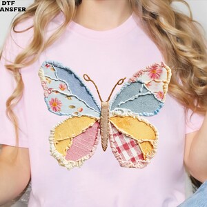 May include: A light pink t-shirt featuring a large, colorful butterfly applique. The butterfly has wings made of patchwork fabrics in shades of blue, yellow, pink, and red, with floral and plaid patterns. The text "DTF TRANSFER" is in the top left corner.