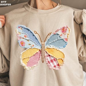 May include: Beige sweatshirt with a patchwork butterfly design. The butterfly is made from various fabrics, including floral, gingham, and solid colours in blue, yellow, pink, and red. The sweatshirt has long sleeves and a crew neck.