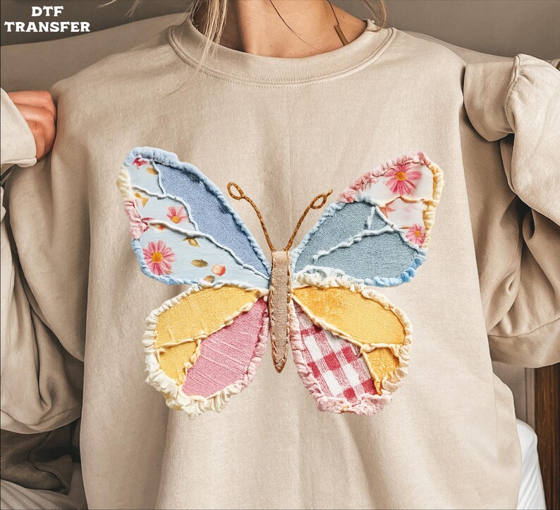 May include: Beige sweatshirt featuring a colorful butterfly applique. The butterfly design incorporates various fabric patterns, including floral, gingham, and solid colors in blue, yellow, pink, and red. The sweatshirt has long sleeves and a crew neck.