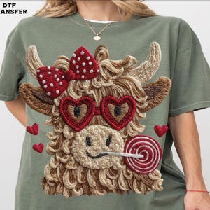 Pode incluir: Camiseta verde-oliva com um design de vaca bordada. A vaca tem &oacute;culos de sol em forma de cora&ccedil;&atilde;o, um la&ccedil;o vermelho e um pirulito. Pequenos cora&ccedil;&otilde;es vermelhos est&atilde;o espalhados ao redor do design. O texto "DTF TRANSFER" est&aacute; no canto superior esquerdo.