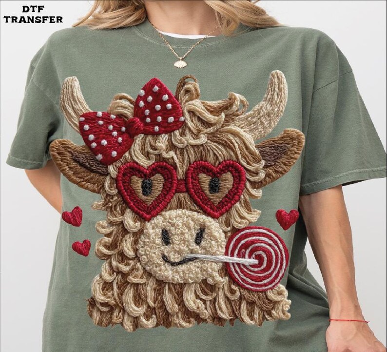 Pode incluir: Camiseta verde-oliva com um design de vaca bordada. A vaca tem &oacute;culos de sol em forma de cora&ccedil;&atilde;o, um la&ccedil;o vermelho e um pirulito. Pequenos cora&ccedil;&otilde;es vermelhos est&atilde;o espalhados ao redor do design. O texto "DTF TRANSFER" est&aacute; no canto superior esquerdo.