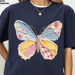 May include: Navy blue t-shirt featuring a large, colorful butterfly applique. The butterfly wings are made of various patterned fabrics, including floral, gingham, and denim. The shirt has a classic crew neck and short sleeves.
