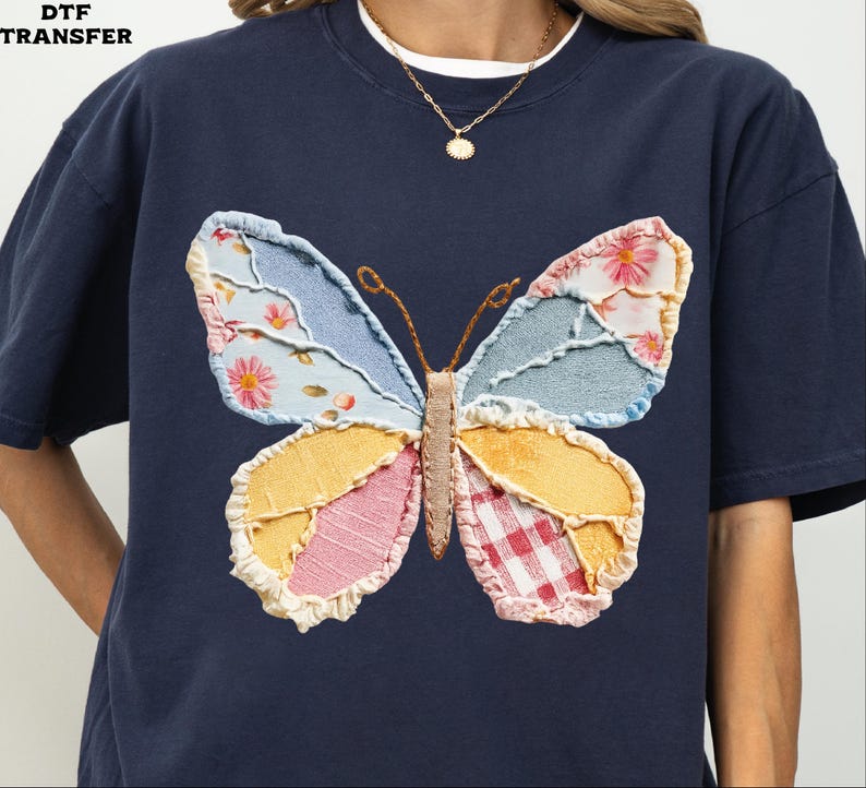 May include: Navy blue t-shirt featuring a large, colorful butterfly applique. The butterfly wings are made of various patterned fabrics, including floral, gingham, and denim. The shirt has a classic crew neck and short sleeves.