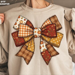 May include: A beige crewneck sweatshirt featuring a large, central graphic of a bow made from patchwork fabric patterns. The design includes orange gingham, brown plaid, white polka dots on mustard, and red floral prints with small pumpkins.