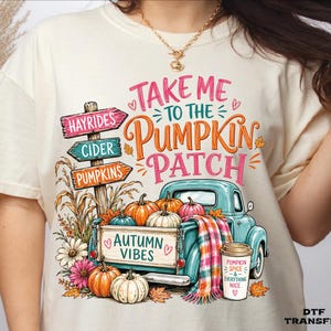 May include: A cream-colored t-shirt featuring a graphic of a teal pickup truck, pumpkins, and a signpost. The text reads: Take Me To The Pumpkin Patch, Autumn Vibes, and Pumpkin Spice & Everything Nice. The design includes colorful fall foliage.