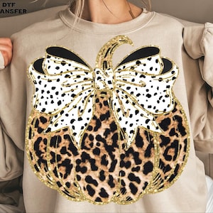 May include: A beige sweatshirt featuring a pumpkin design. The pumpkin has a leopard print body, a gold glitter outline, and a large bow with a black and white dalmatian print. The stem and bow details are outlined in gold glitter.