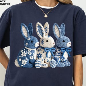 May include: Navy blue t-shirt featuring three crocheted bunny rabbits. The bunnies have blue and white floral patterns, with blue bows and ears. The text "DTF TRANSFER" is in the upper left corner.