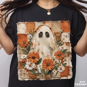 May include: A black t-shirt featuring a square patch design with a central ghost figure surrounded by orange and white flowers. The background of the patch consists of a checkered pattern in shades of beige and orange with visible stitching details.