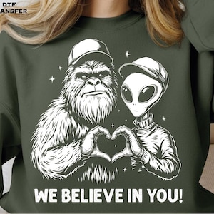 May include: Dark green sweatshirt with a white graphic of a Bigfoot and an alien forming a heart with their hands. The text "WE BELIEVE IN YOU!" is below the graphic. The sweatshirt has long sleeves.