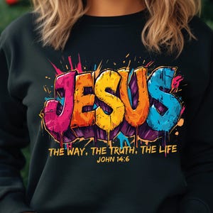May include: Black sweatshirt featuring the word "JESUS" in colorful graffiti-style lettering. The design includes the text "THE WAY, THE TRUTH, THE LIFE JOHN 14:6" below the word. The sweatshirt has a crew neck and long sleeves.