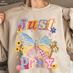 May include: Beige sweatshirt with a colorful butterfly and the words "Just Pray." The butterfly has blue, yellow, pink, and plaid wings. Yellow sunflowers and a cross with flowers are also featured in the design.