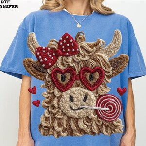 Pode incluir: Camiseta azul claro com um design bordado de vaca das Terras Altas. A vaca tem &oacute;culos de sol em forma de cora&ccedil;&atilde;o, um la&ccedil;o vermelho e um pirulito. Pequenos cora&ccedil;&otilde;es vermelhos est&atilde;o espalhados ao redor do design. O texto "DTF TRANSFER" est&aacute; no canto superior esquerdo.