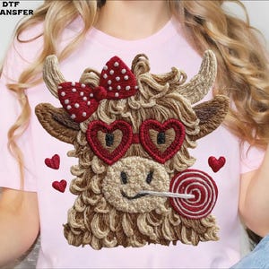 Pode incluir: Camiseta rosa com um design bordado de vaca das Terras Altas. A vaca tem &oacute;culos de sol em forma de cora&ccedil;&atilde;o, um la&ccedil;o vermelho com bolinhas brancas e um pirulito. Pequenos cora&ccedil;&otilde;es vermelhos est&atilde;o espalhados ao redor da vaca.