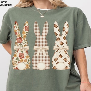 May include: Sage green t-shirt featuring three bunny designs. The bunnies have floral, gingham, and daisy patterns, with cream-colored bows and fluffy white tails. The text "DTF TRANSFER" is in the upper left corner.