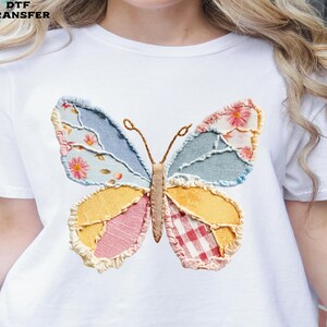May include: White t-shirt featuring a colorful butterfly design. The butterfly has patchwork wings in shades of blue, yellow, pink, and red, with floral and gingham patterns. The text "DTF TRANSFER" is in the upper left corner.